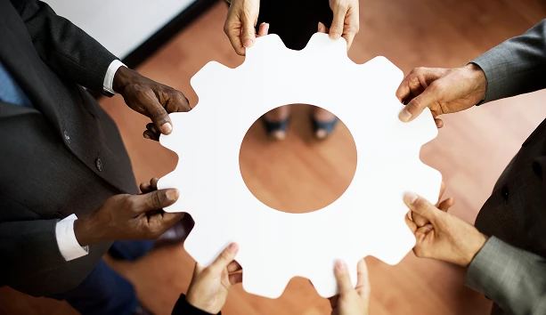 Diverse business professionals holding a large gear together, symbolizing teamwork, collaboration, process improvement, and organizational success.
